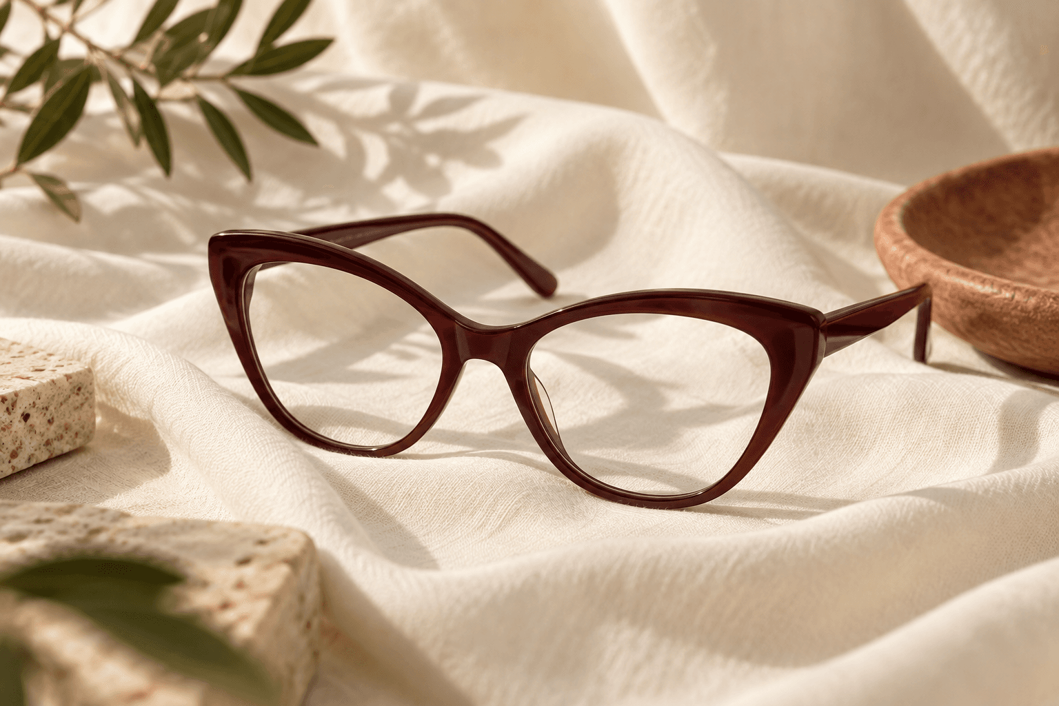 Dark cherry cat-eye glasses resting on ivory fabric beside a soft leaf shadow.